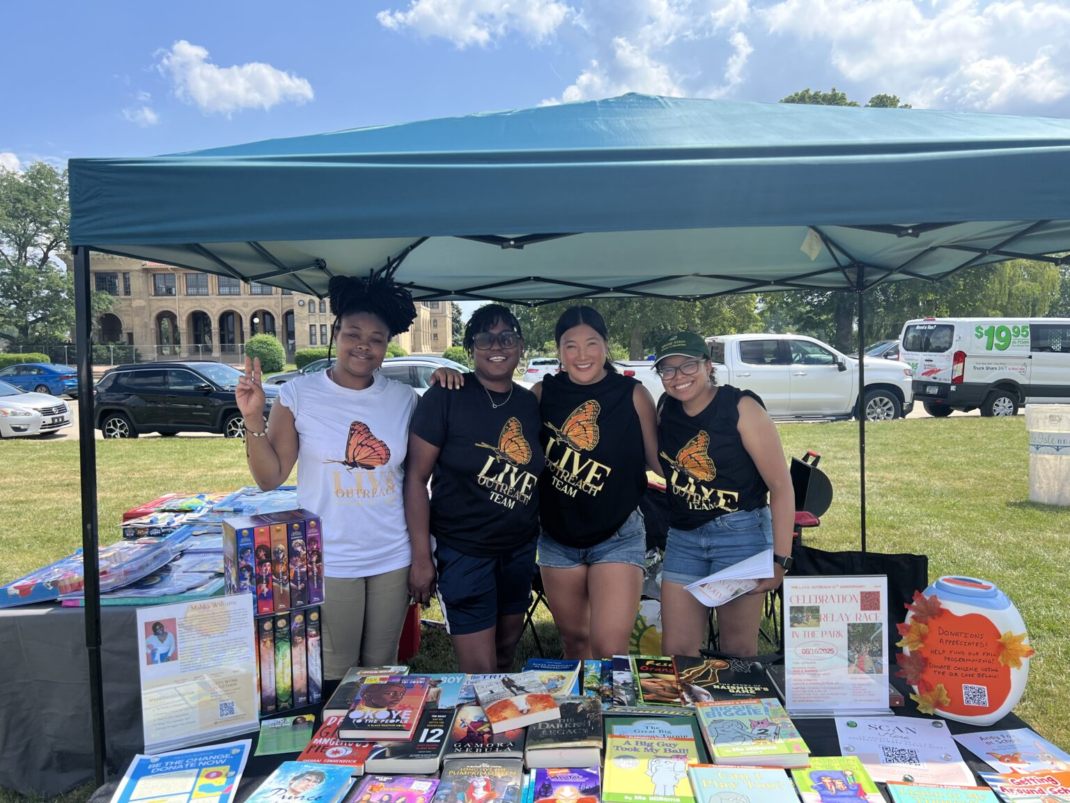 Four L.I.V.E. Outreach team members in butterfly logo shirts smiling behind a table of children's books at an outdoor community event