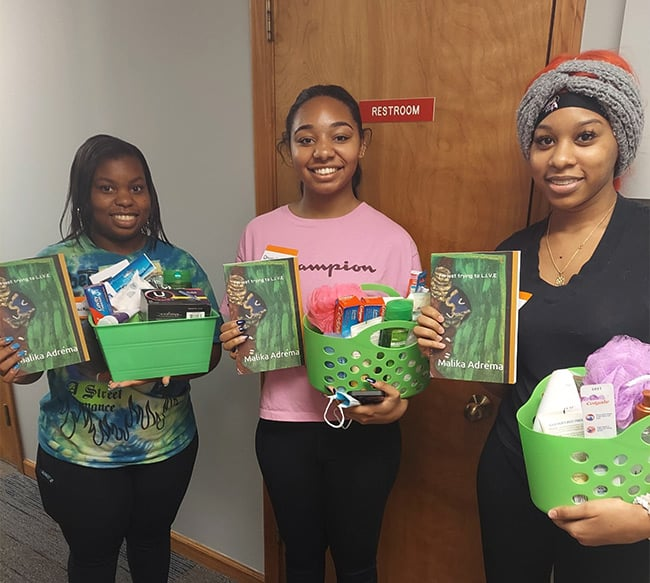 Three young women smiling and holding baskets filled with toiletries and copies of "I'm just trying to L.I.V.E." by Malika Adrema