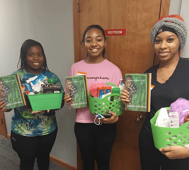 Three young women smiling and holding baskets filled with toiletries and copies of "I'm just trying to L.I.V.E." by Malika Adrema